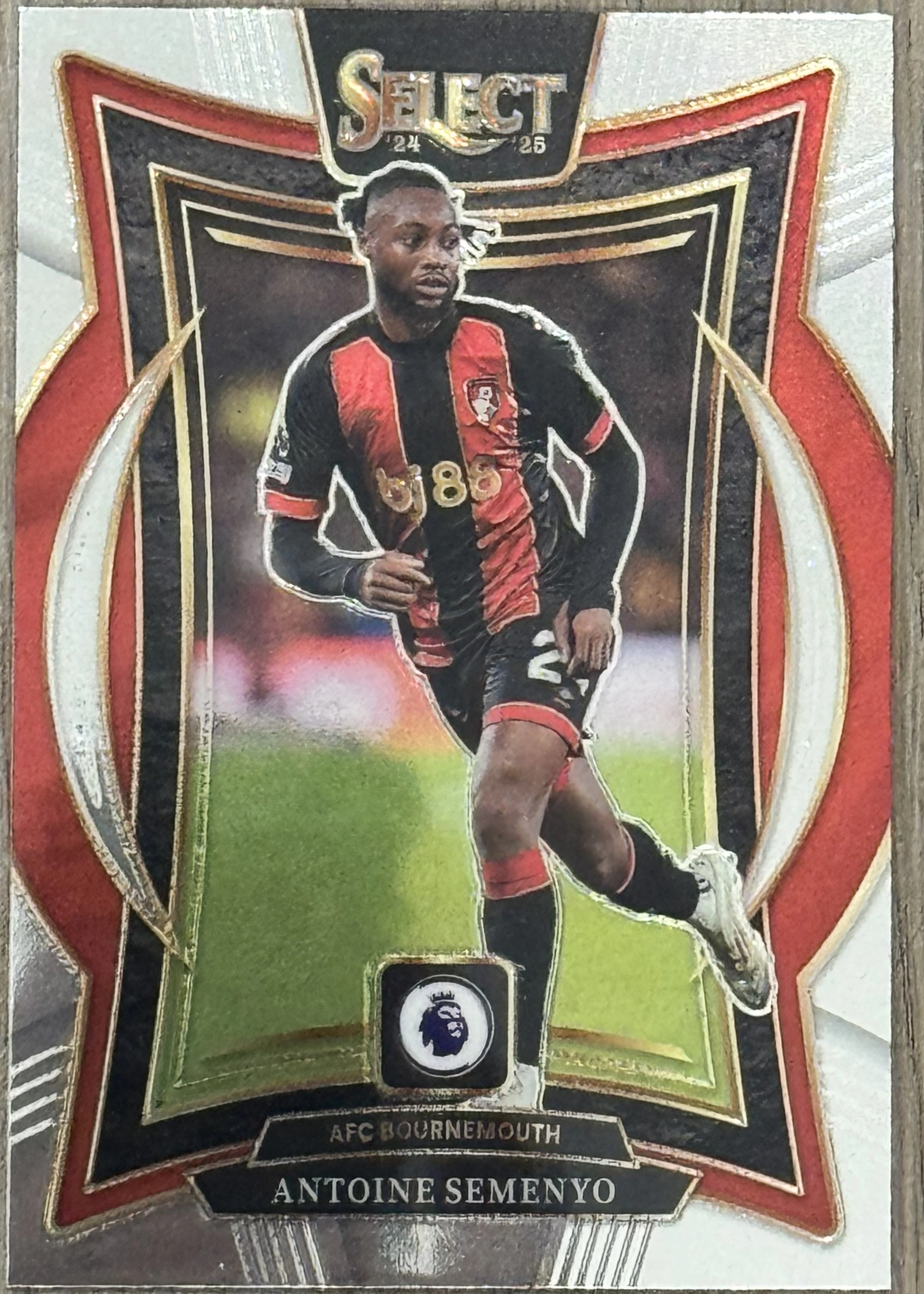 Front of 2024-25 Panini Select Premier League Soccer Antoine Semenyo Terrace Silver Prizm, a soccer trading card featuring a player in a black and red striped jersey with the number 85, set against a white and silver background with a red…