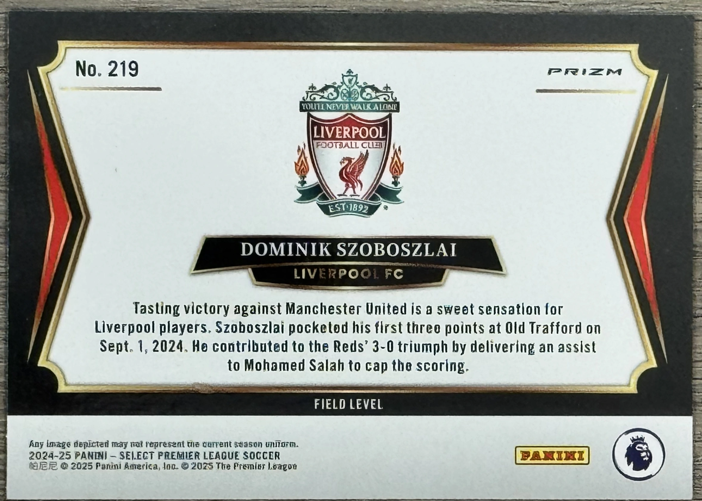 Back of 2024-25 Panini Select Premier League Soccer Dominik Szoboszlai #219 2024-25 Select Premier League Soccer #219 Dominik Szoboszlai Red Prizm, the back of a soccer trading card with a white central panel framed by a black border with red and gold foil accents. The card is rotated 90 degrees…
