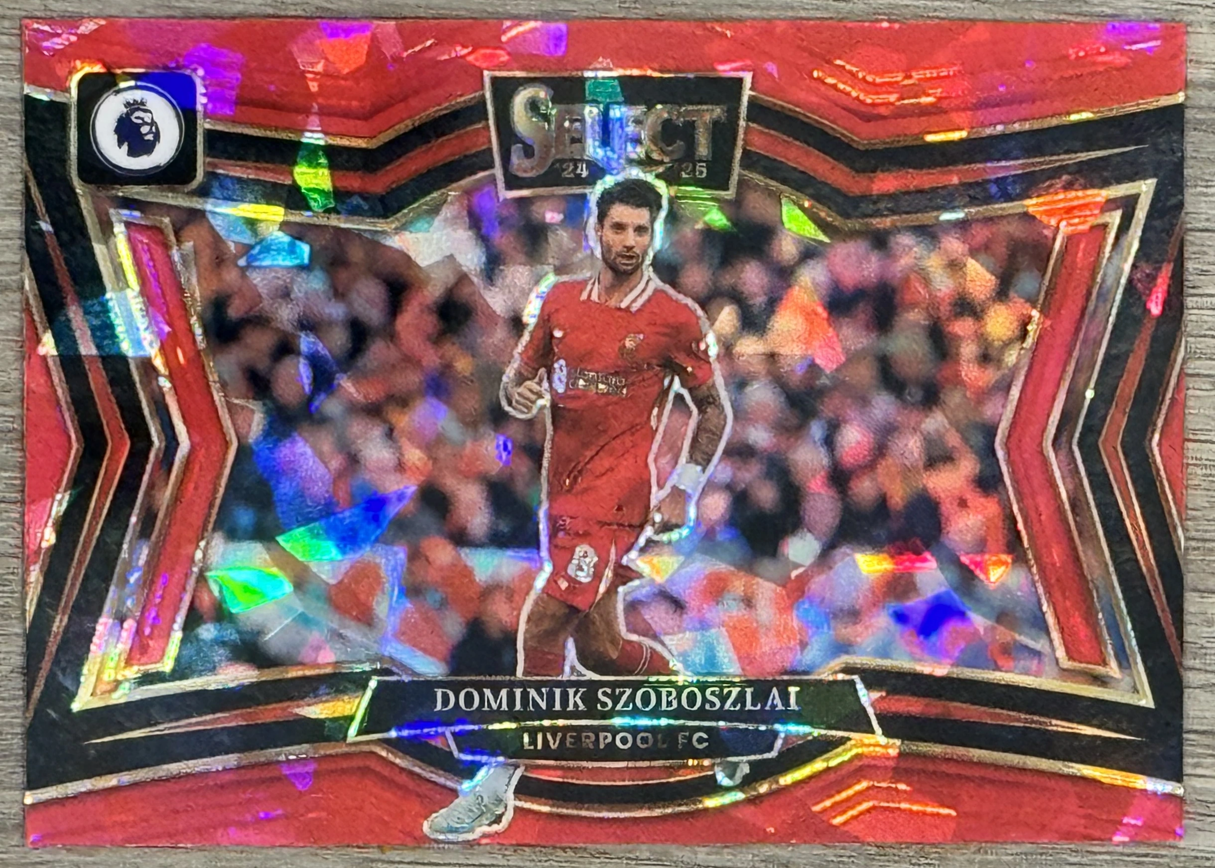 Front of 2024-25 Panini Select Premier League Soccer Dominik Szoboszlai #219 2024-25 Select Premier League Soccer #219 Dominik Szoboszlai Red Prizm, a soccer trading card with a red and black chevron border and a holographic prizm background. The central image shows a player in a red Liverpool…
