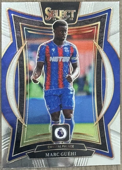 Front of 2024-25 Panini Select Premier League Soccer Marc Guéhi Terrace Blue Prizm, a soccer trading card featuring a full-body die-cut image of a player in a blue and red striped Crystal Palace jersey with 'NET88' sponsor, standing…