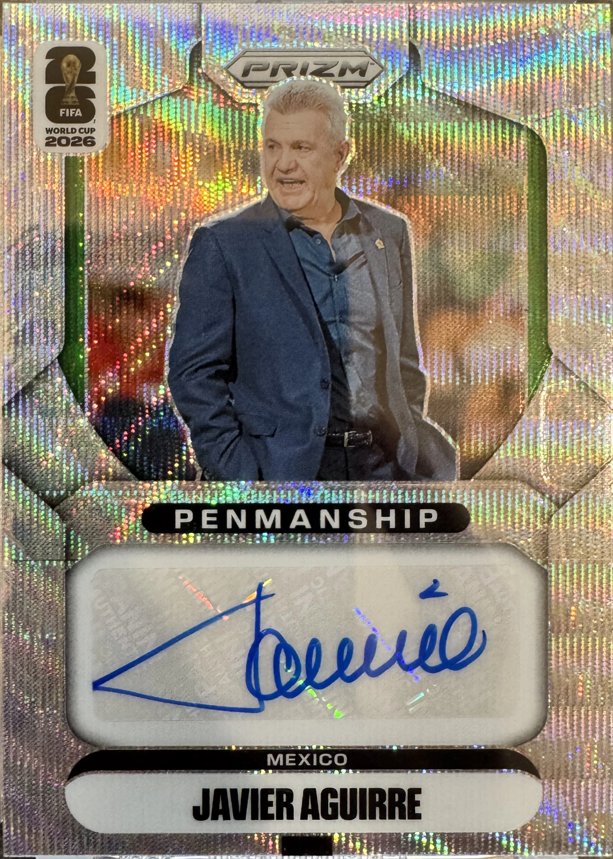 Front of 2026 Panini 2026 Panini Prizm FIFA World Cup 2026 Soccer Javier Aguirre #P-JA Penmanship Prizm autographed, a vertical collectible card with a holographic, refractor-style background featuring vertical rainbow stripes. The top left displays a '26 FIFA…